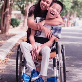 Teen boy using a wheelchair with his mothers arms wrapped around him, both smiling with joy.