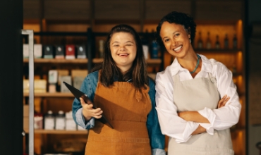 A young adult woman with Down Syndrome, standing beside another smiling woman with a chef apron.
